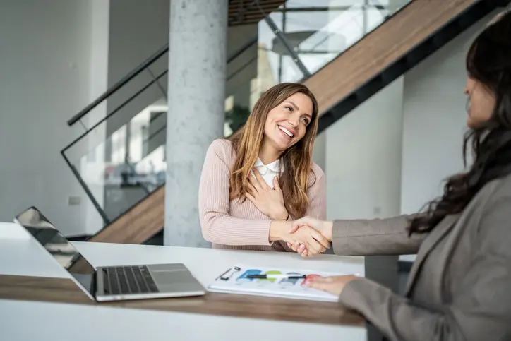 Happy businesswoman receiving good news, smiling cheerfully while shaking hands with a new client after a successful meeting in a modern office, symbolizing agreement and partnership
